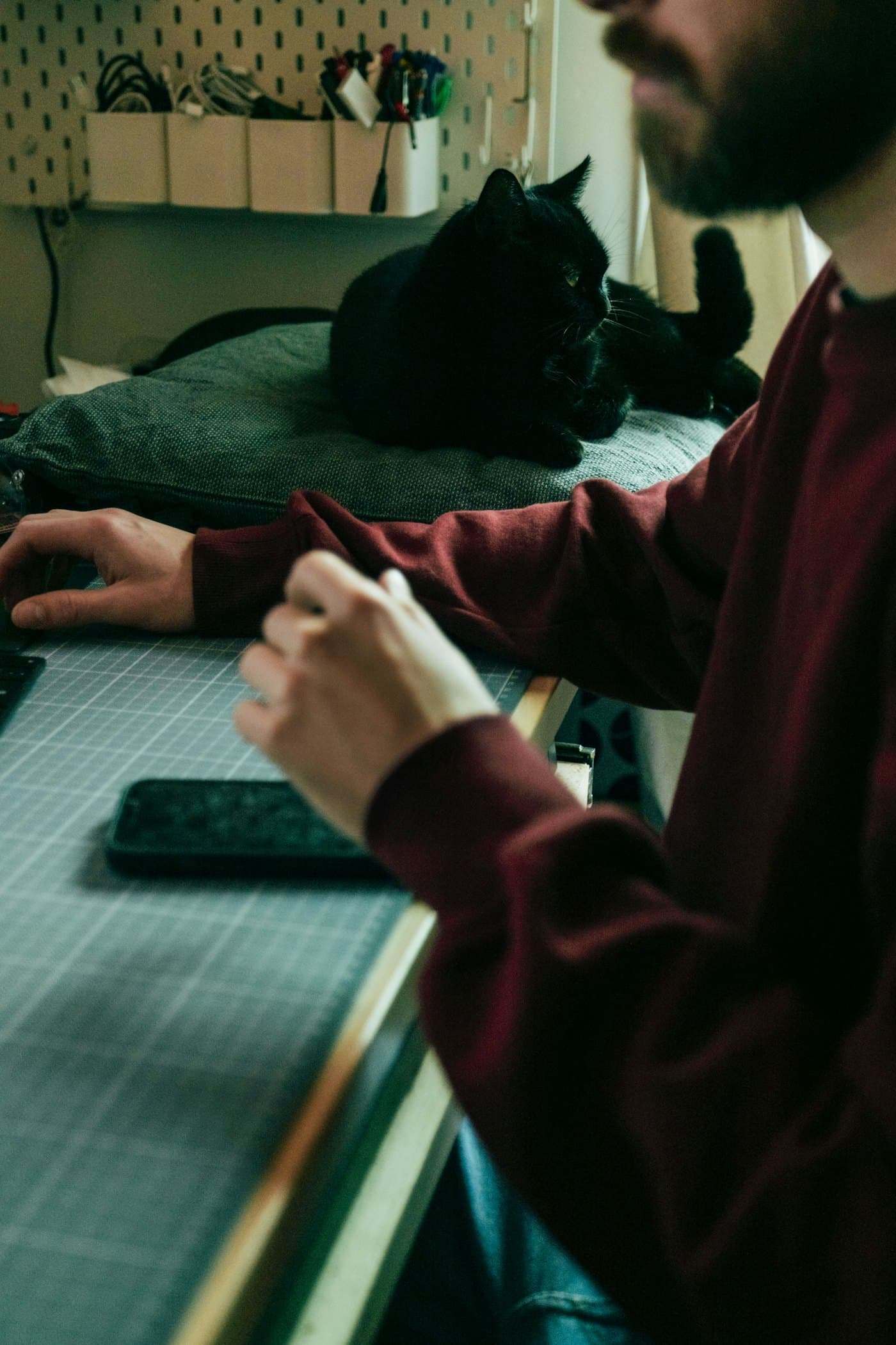Maker aan een werktafel thuis met snijmat en telefoon, een kat kijkt toe op de achtergrond